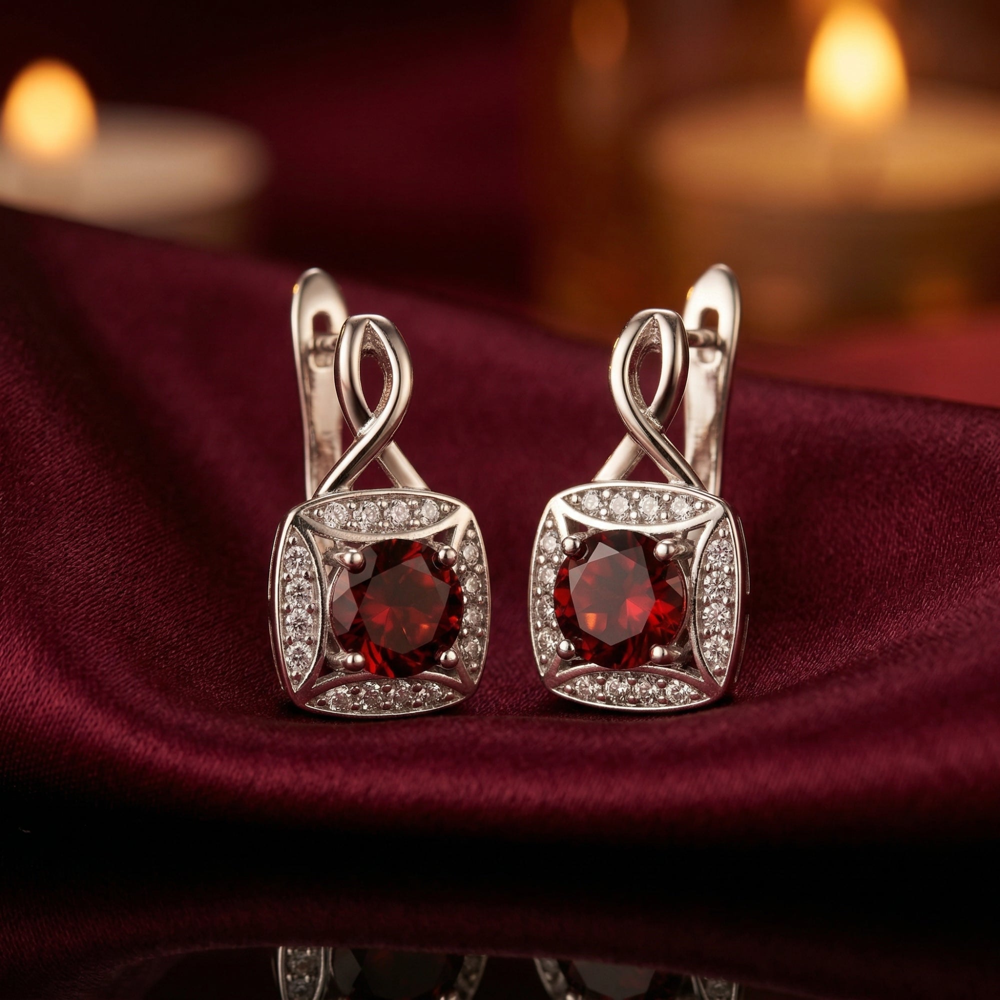 Pair of round-cut garnet drop earrings in sterling silver with zircon halos on a dark red fabric background.