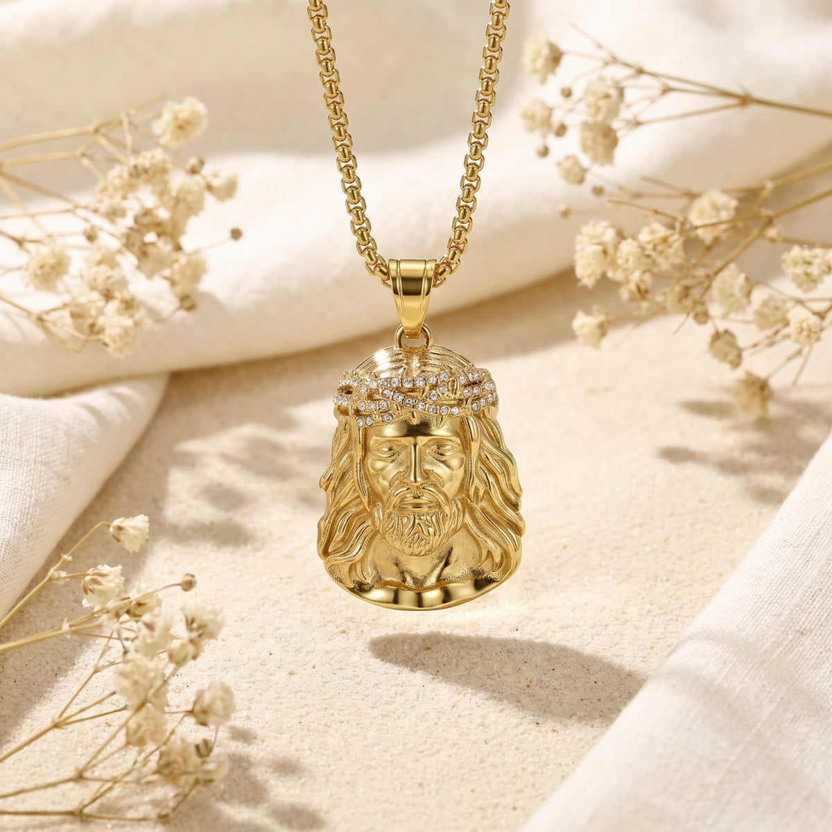 Front-facing gold-finish Jesus portrait pendant suspended above sand with dried flowers.