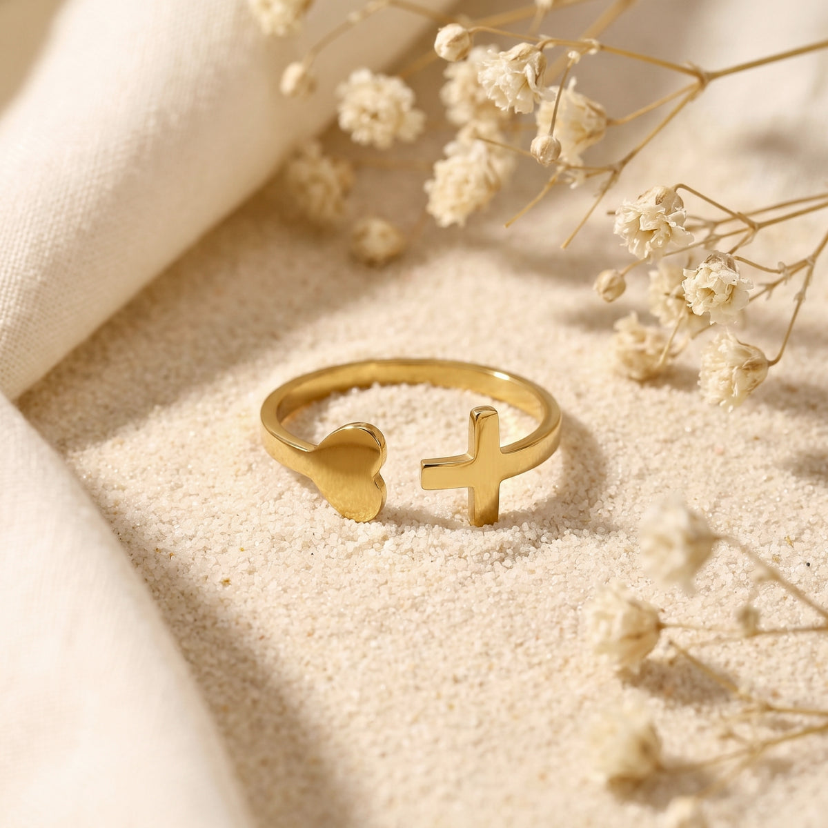 Gold-tone open ring with heart and cross ends lying on sand among dried white flowers.