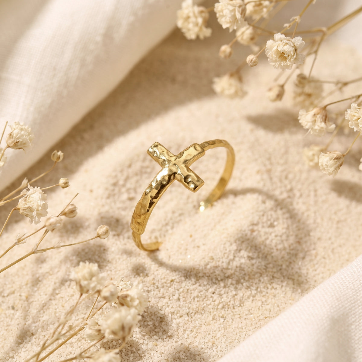 Angled gold-tone hammered cross ring on sand, showing its open band and textured surface.