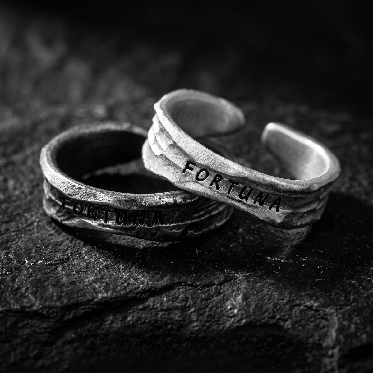 Close-up of two textured open silver rings overlapping on a dark textured surface, both stamped FORTUNA.