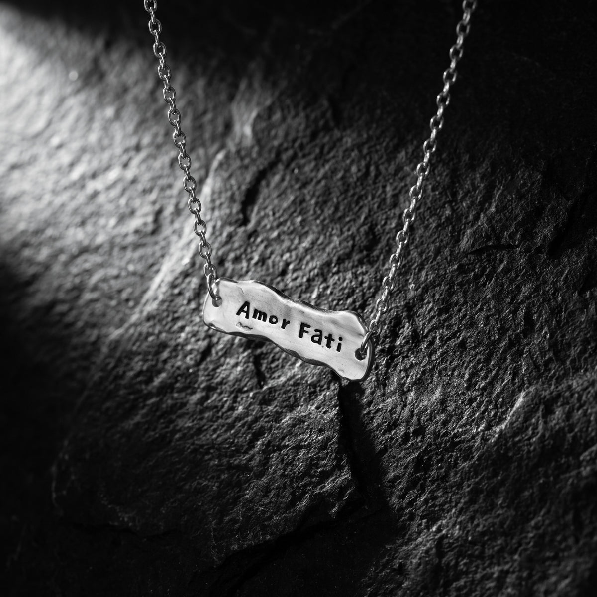Close-up of the Amor Fati bar necklace resting on textured dark rock, with the inscription facing up.