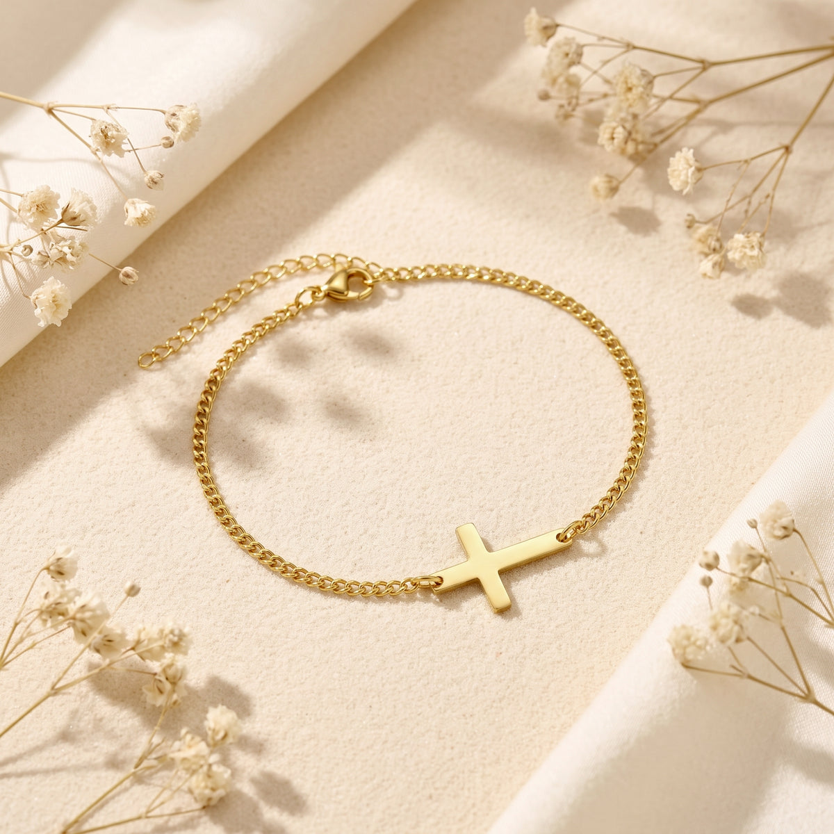 Overhead view of a gold-toned horizontal cross bracelet arranged in a circle on sand with dried flowers.
