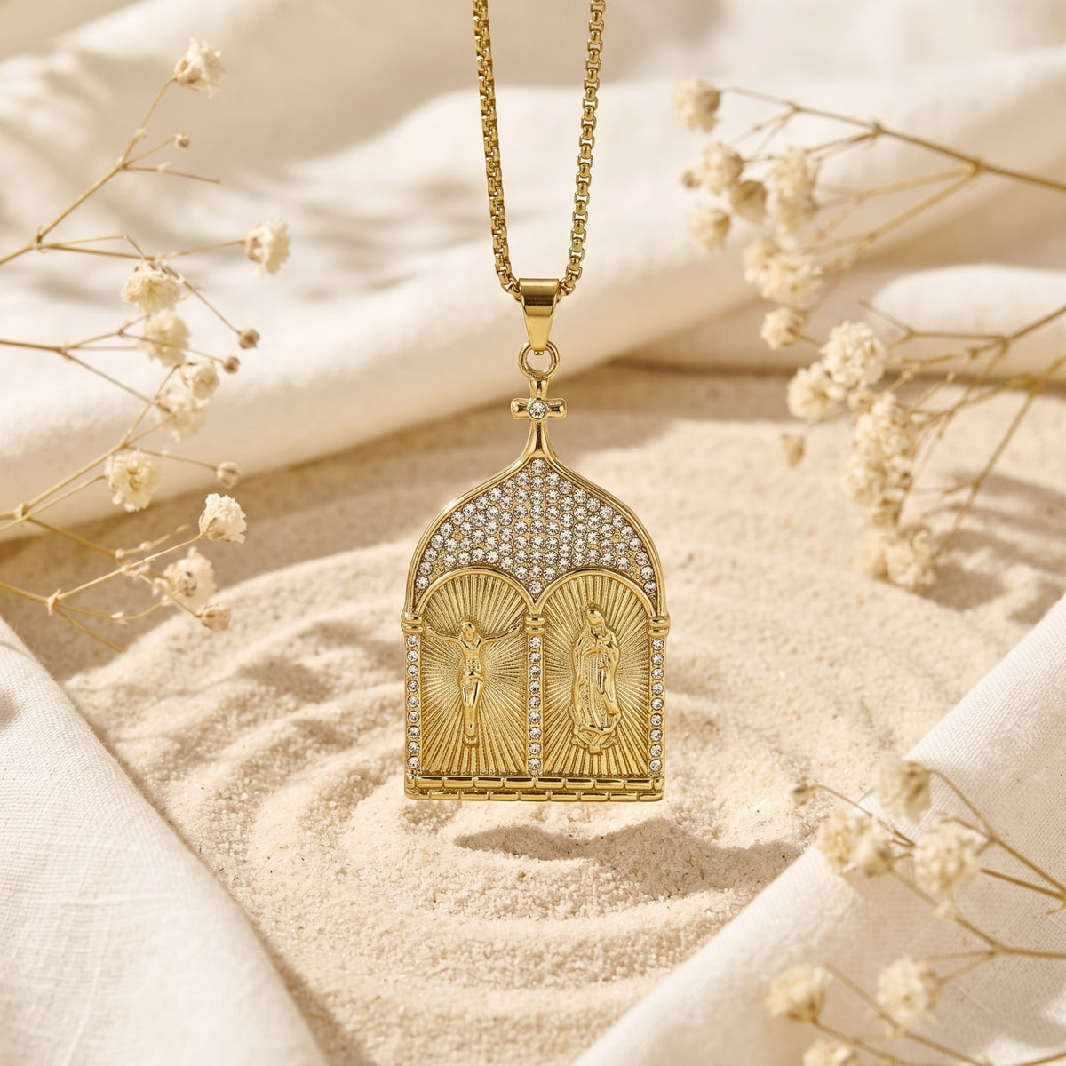 Gold-tone diptych pendant suspended upright above sand with dried white flowers.