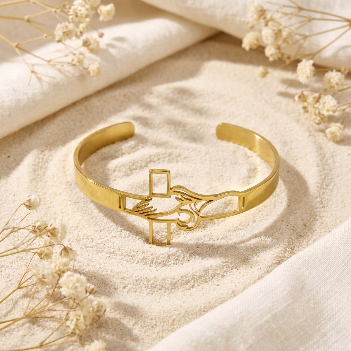 Front view of the gold-tone open cuff bracelet with praying hands and cross on sand.