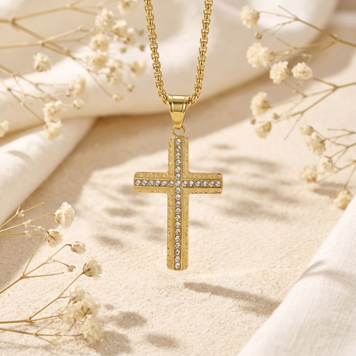 Front view of the gold-tone cross pendant with cubic zirconia accents suspended above a sand backdrop.