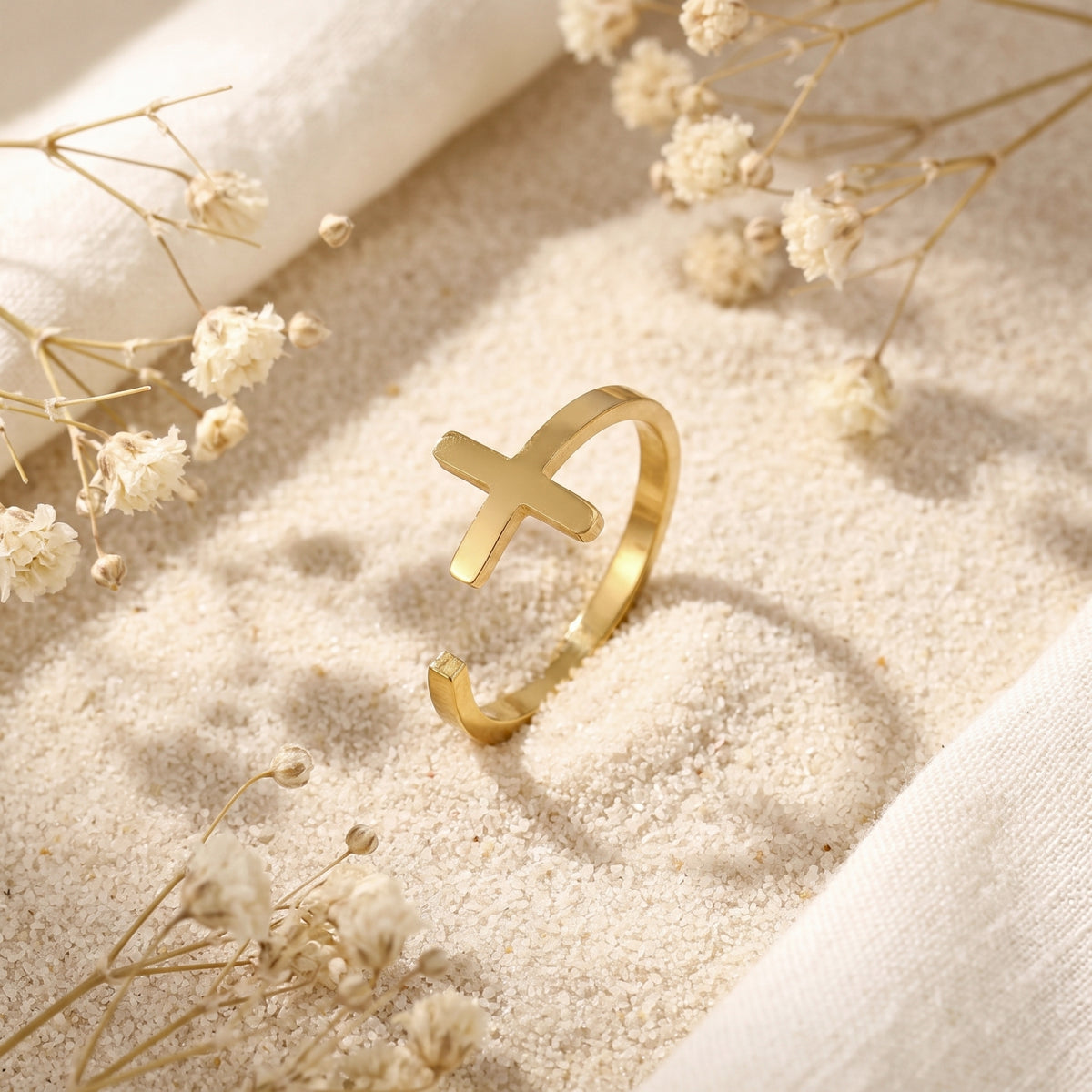 Angled view of the gold-tone cross open ring standing in white sand near dried flowers.