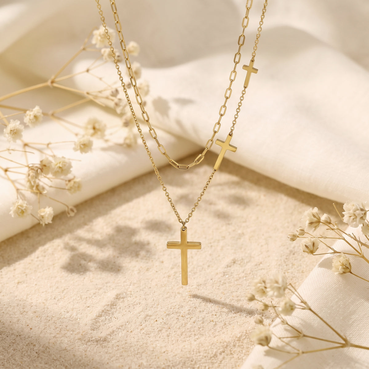 Centered view of the gold-tone layered cross necklace displayed above sand with folded white fabric and dried flowers.