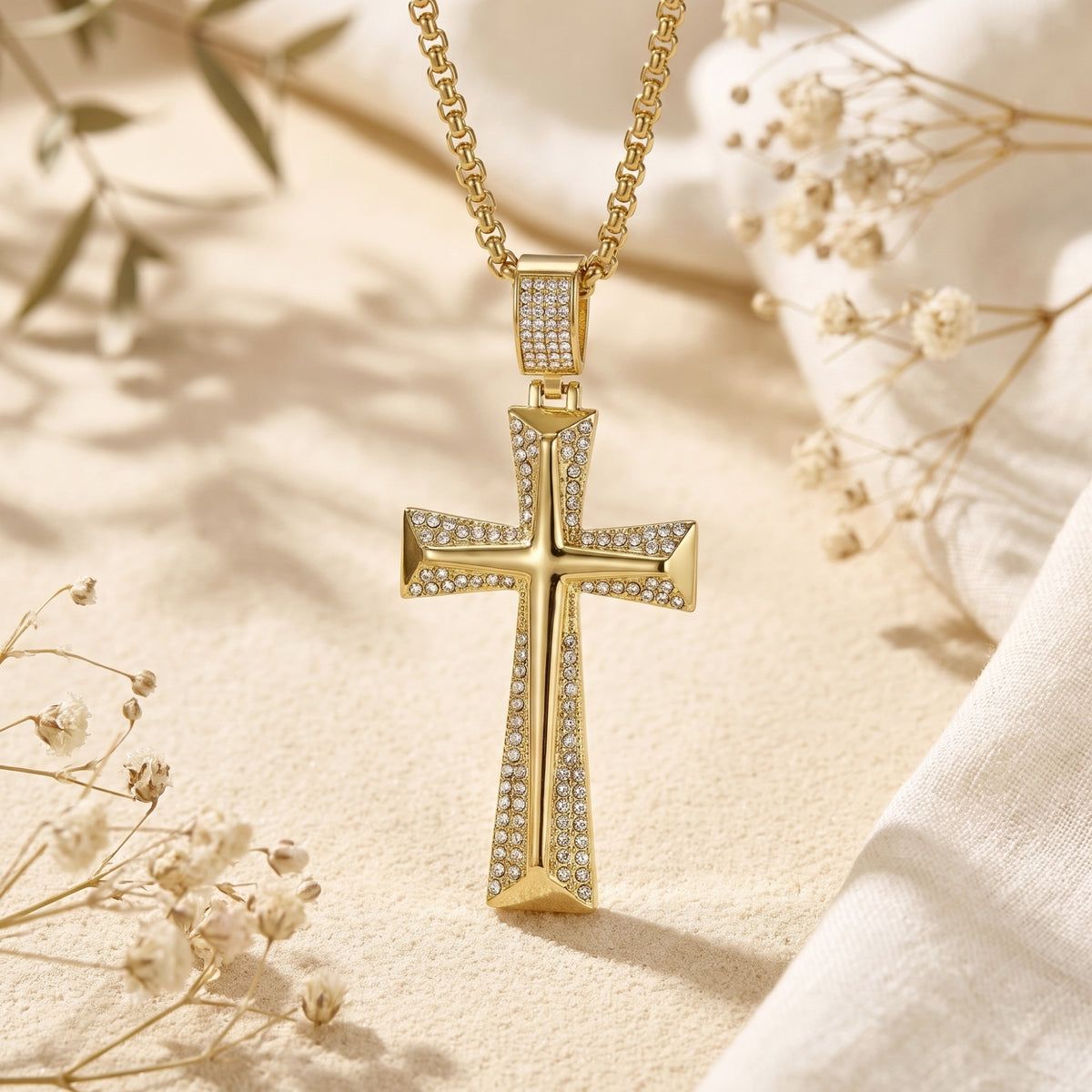 Front-facing gold-tone beveled cross pendant with pavé accents, suspended over sand and dried flowers.