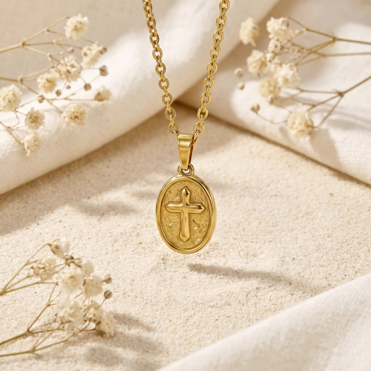 Gold-tone oval cross medallion pendant hanging over sand, linen, and dried white flowers.