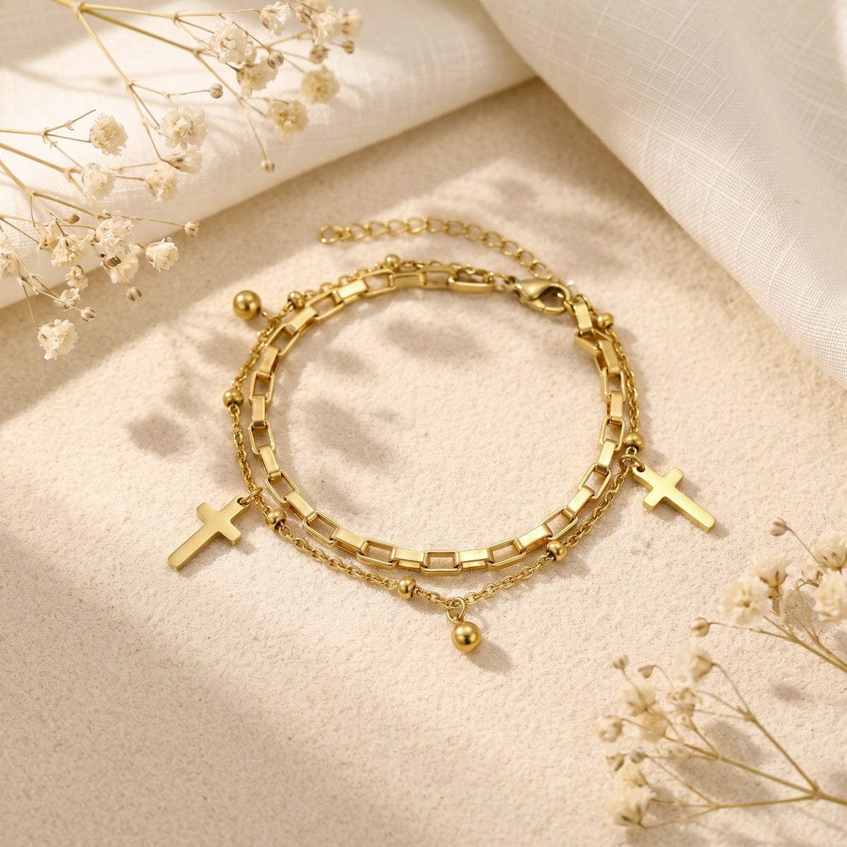 Angled view of the gold-tone double-layered cross bracelet on a textured surface with white cloth and dried flowers.