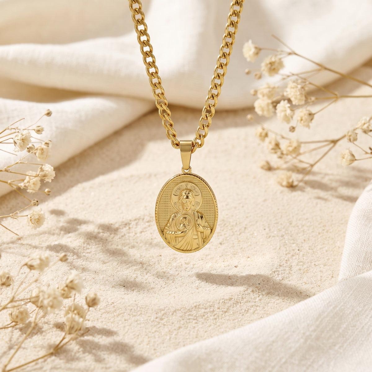 Gold-tone Saint Jude pendant hanging vertically above sand, framed by white fabric and dried flowers.