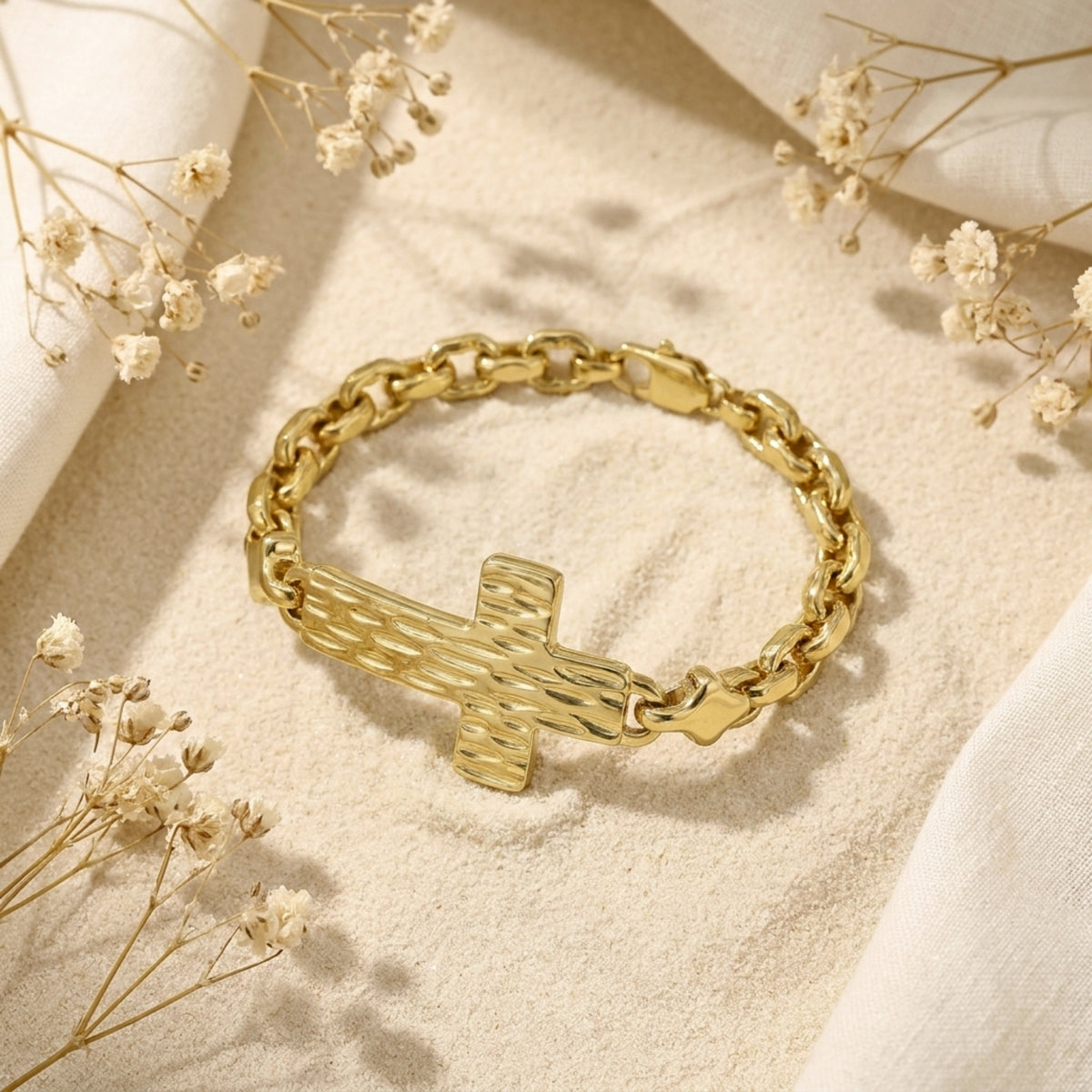 Angled view of a gold-tone textured cross link bracelet resting on sand beside dried flowers, with clasp visible.