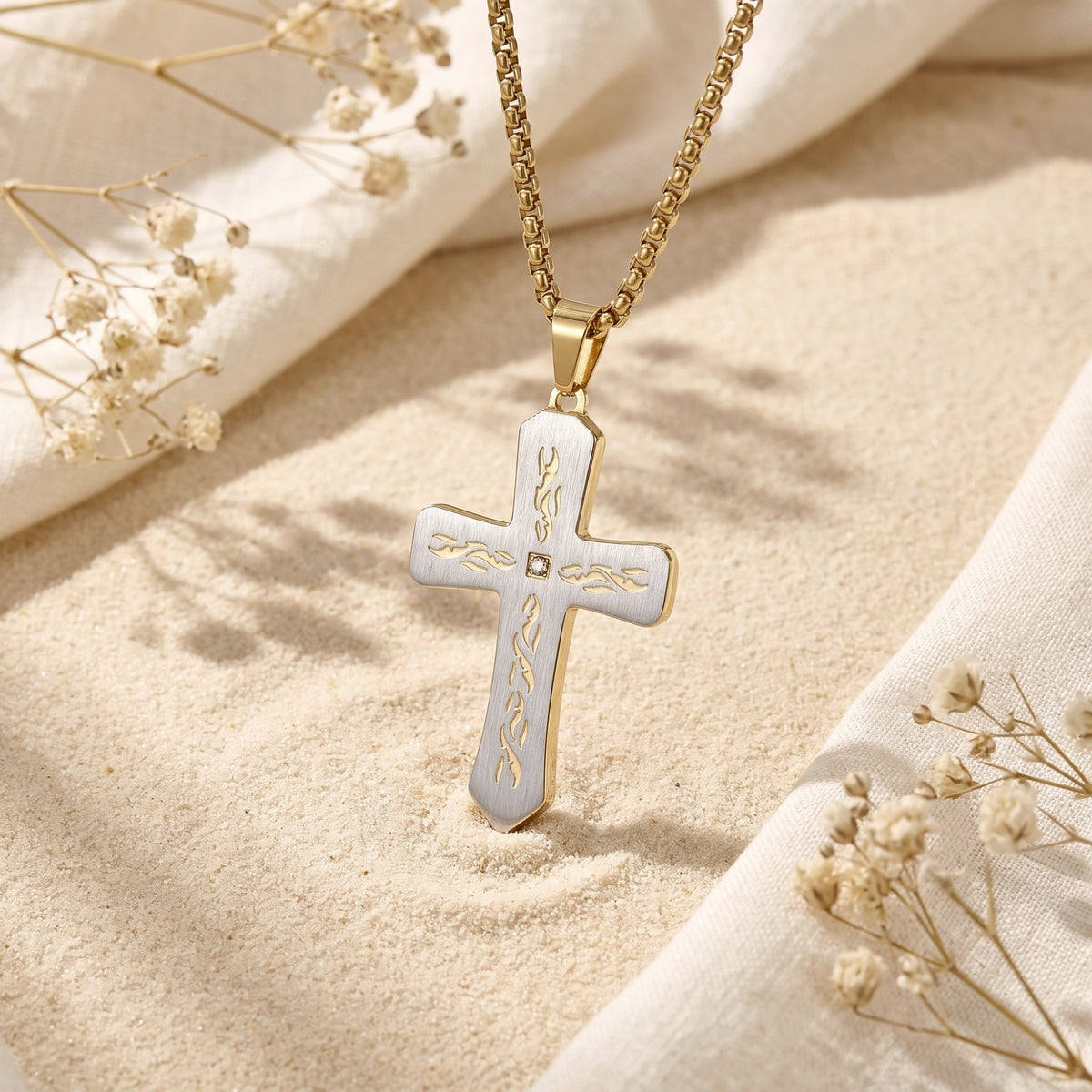 Close-up of a two-tone cross pendant standing upright in sand, showing etched gold-tone accents and a center stone.