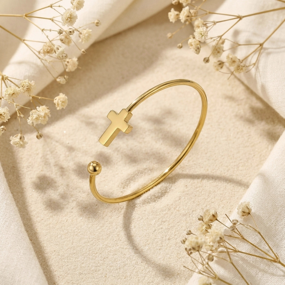 Gold-tone open cuff bracelet tilted on textured sand surrounded by delicate dried floral sprigs.