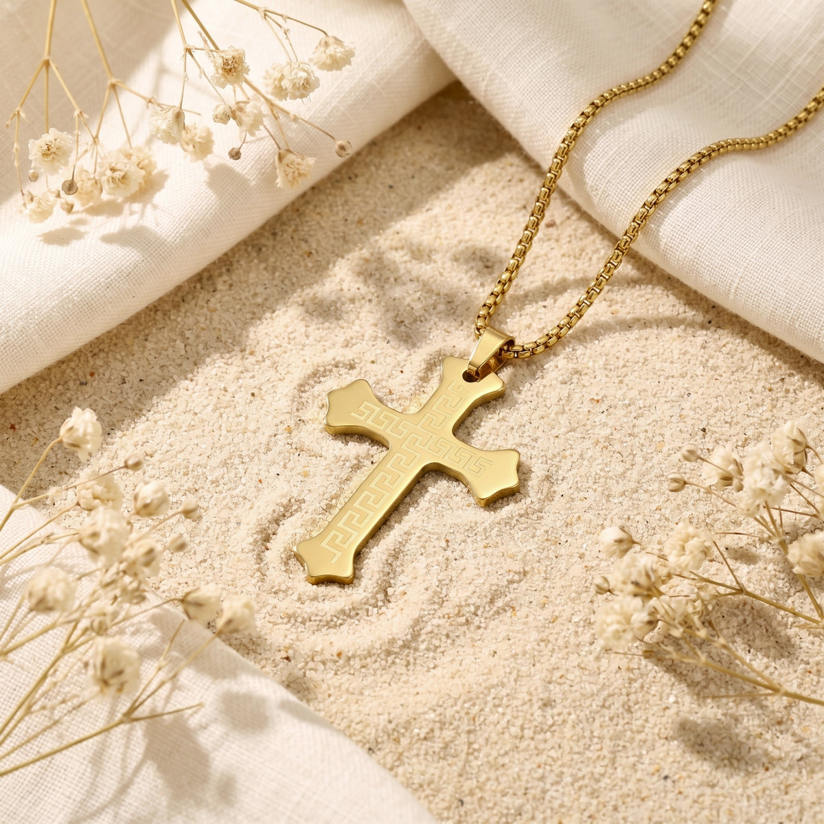 Gold-toned Greek key cross necklace lying flat on sand with white fabric and dried flowers.