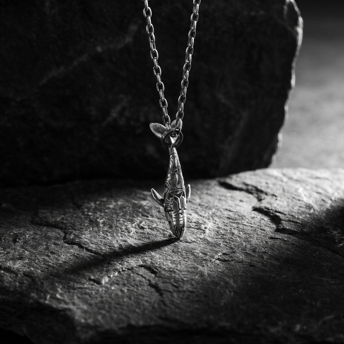 Carved silver shark pendant hanging vertically over a dark slate surface in dramatic lighting.