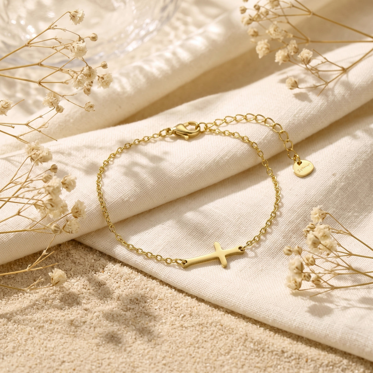 Gold-tone horizontal cross bracelet resting on draped cream linen, with sand and dried flowers around it.