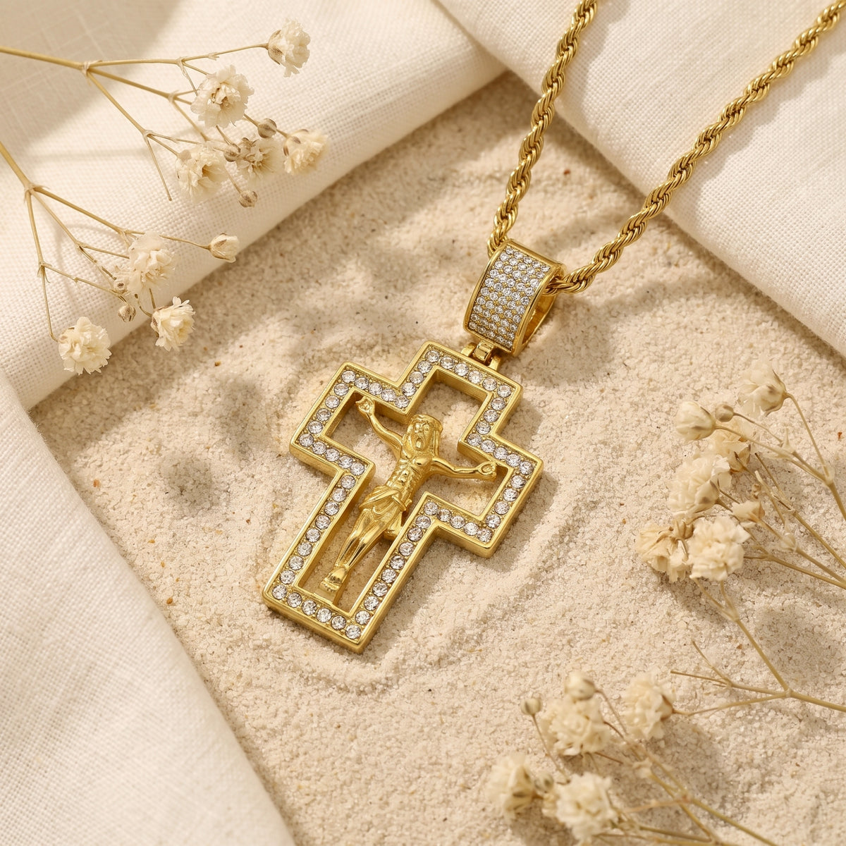 Angled flat lay of a gold-tone cutout crucifix pendant with clear stones on sand beside dried flowers.