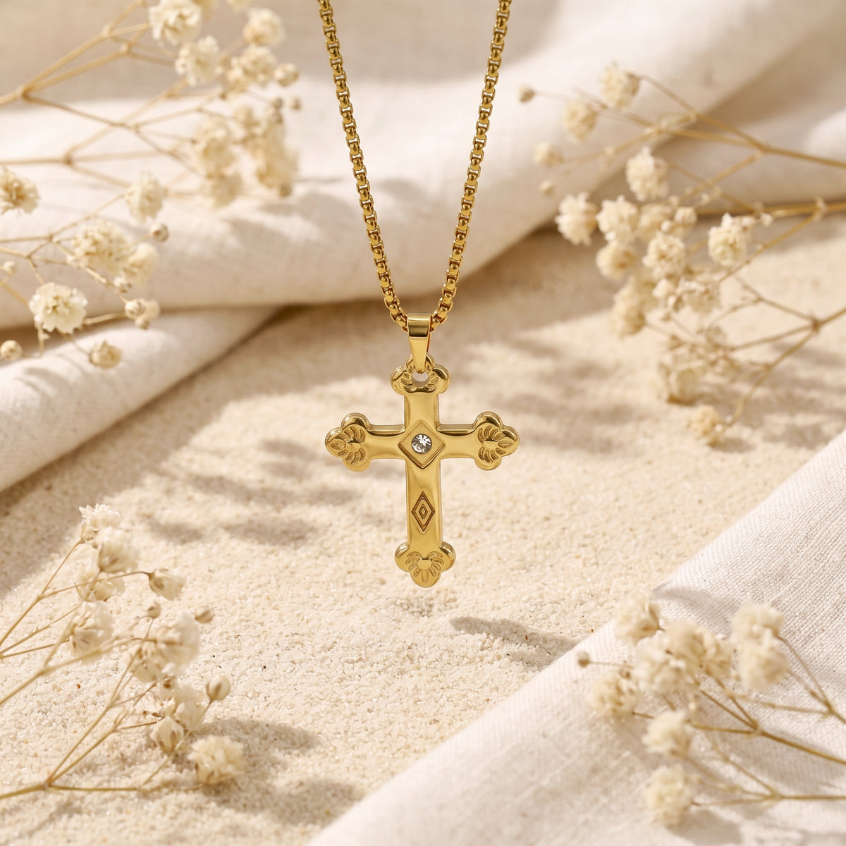 Gold-tone floriated cross necklace suspended over sand with white dried flowers and folded cream fabric.