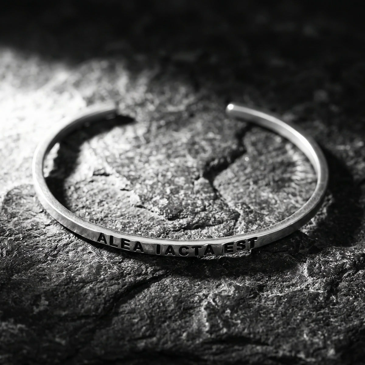 Open silver cuff engraved “ALEA IACTA EST” resting on a textured rock surface.