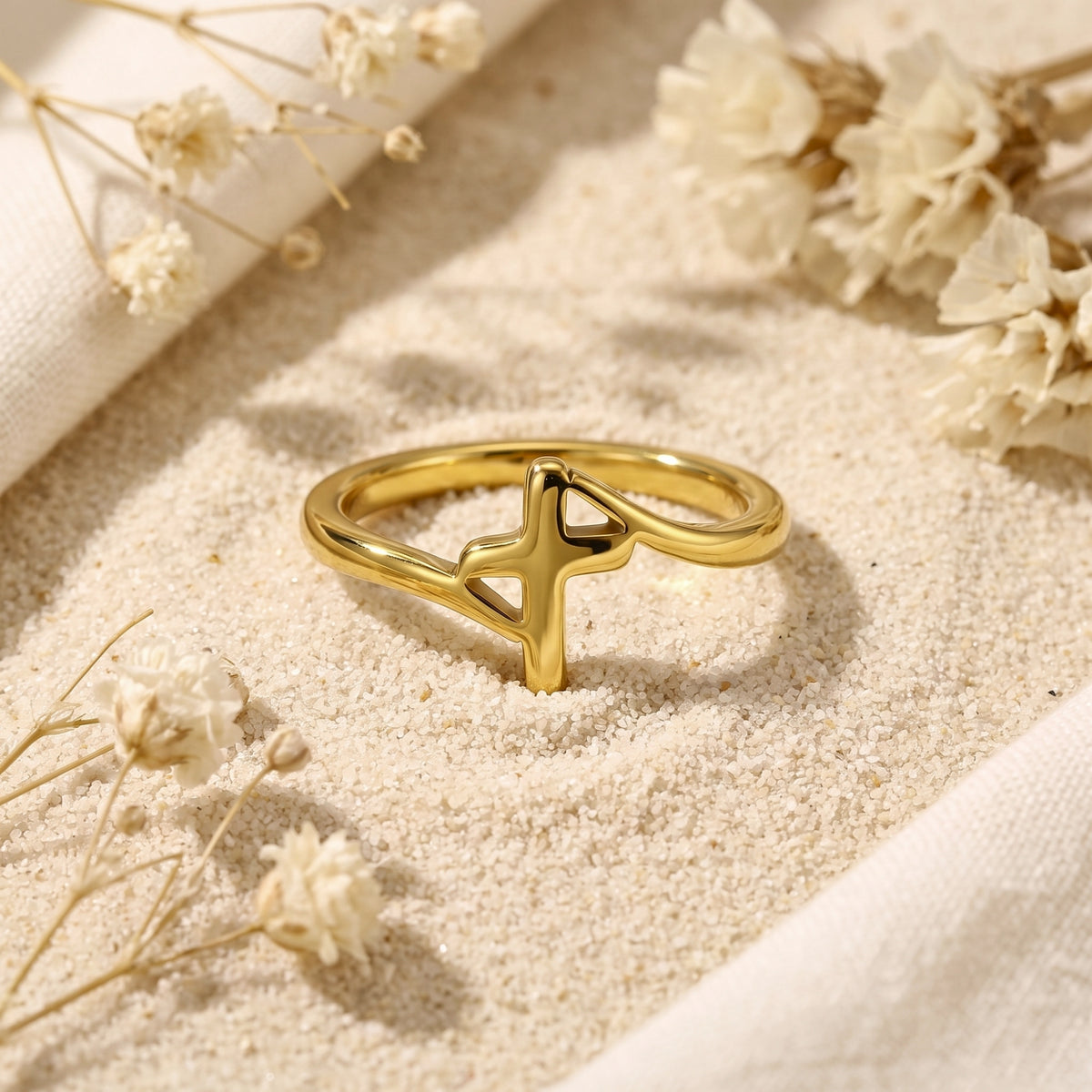 Front-facing gold-tone openwork cross ring standing in fine sand among dried flowers.
