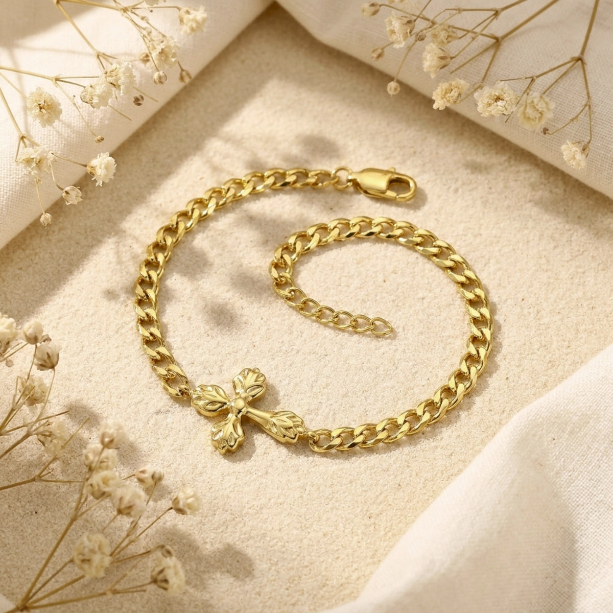Gold-tone curb chain bracelet laid in a loop on sand, with its lobster clasp, extender, and cross centerpiece visible.