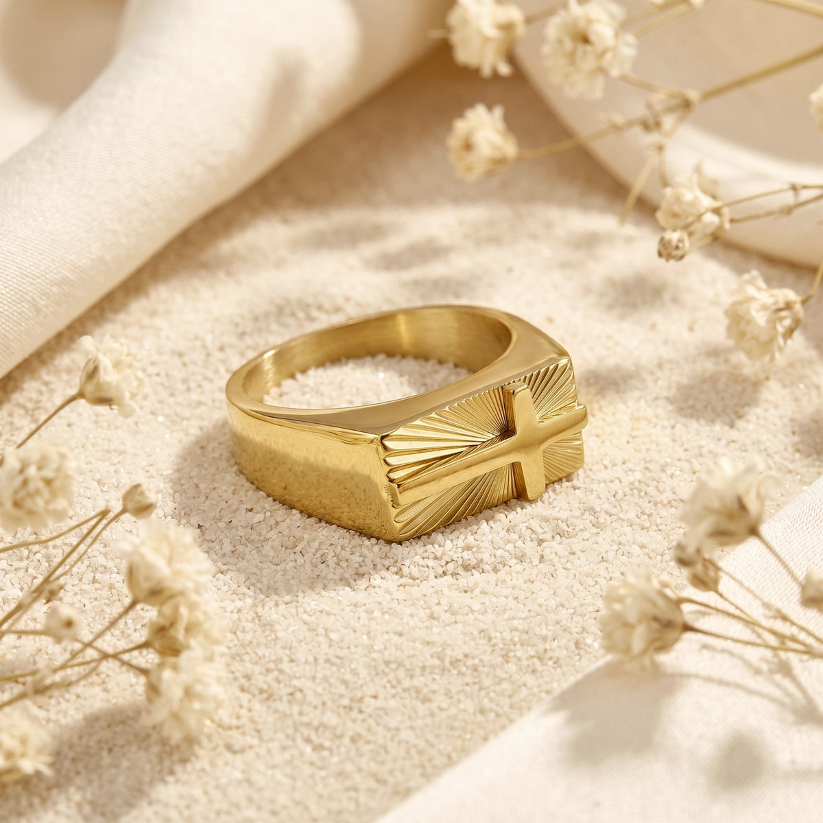 Angled view of the gold-tone sunburst cross signet ring lying on white sand among dried flowers.