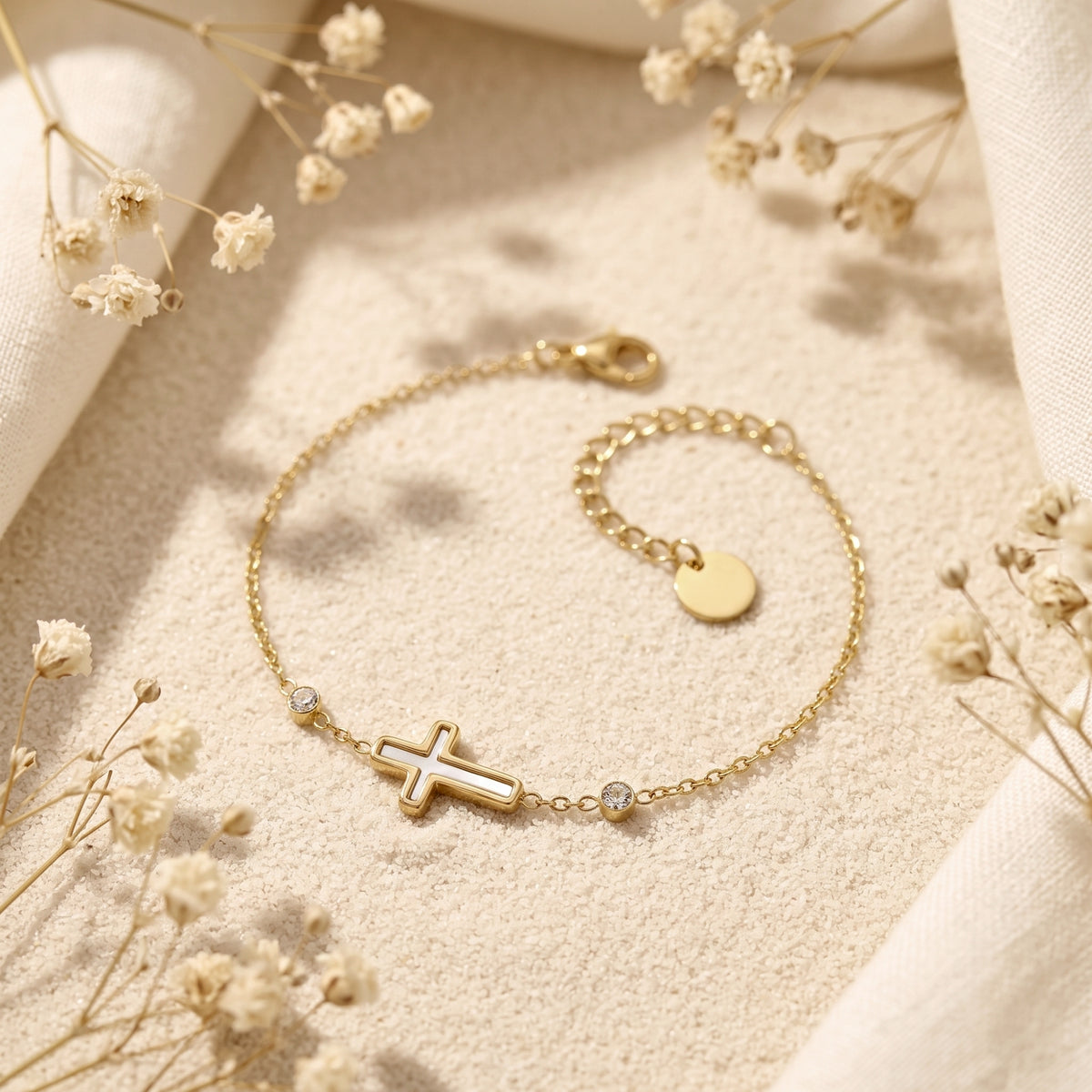 Gold-tone cross bracelet laid in a curve on sand, displaying its chain extension and round disc tag.