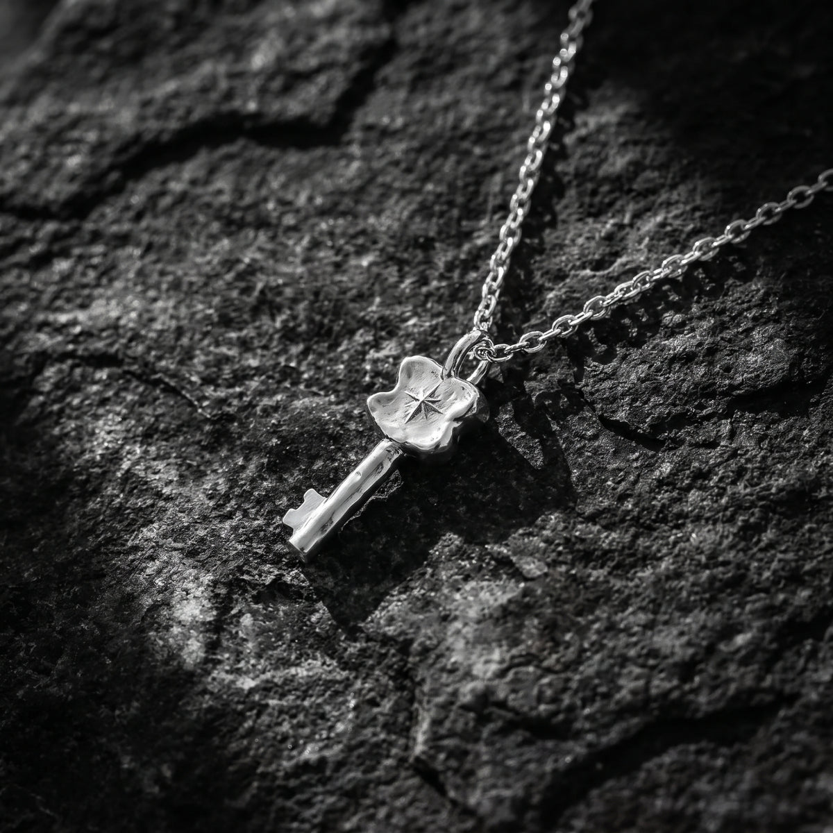 Silver starburst key pendant lying diagonally on textured rock with its chain extending across the surface.