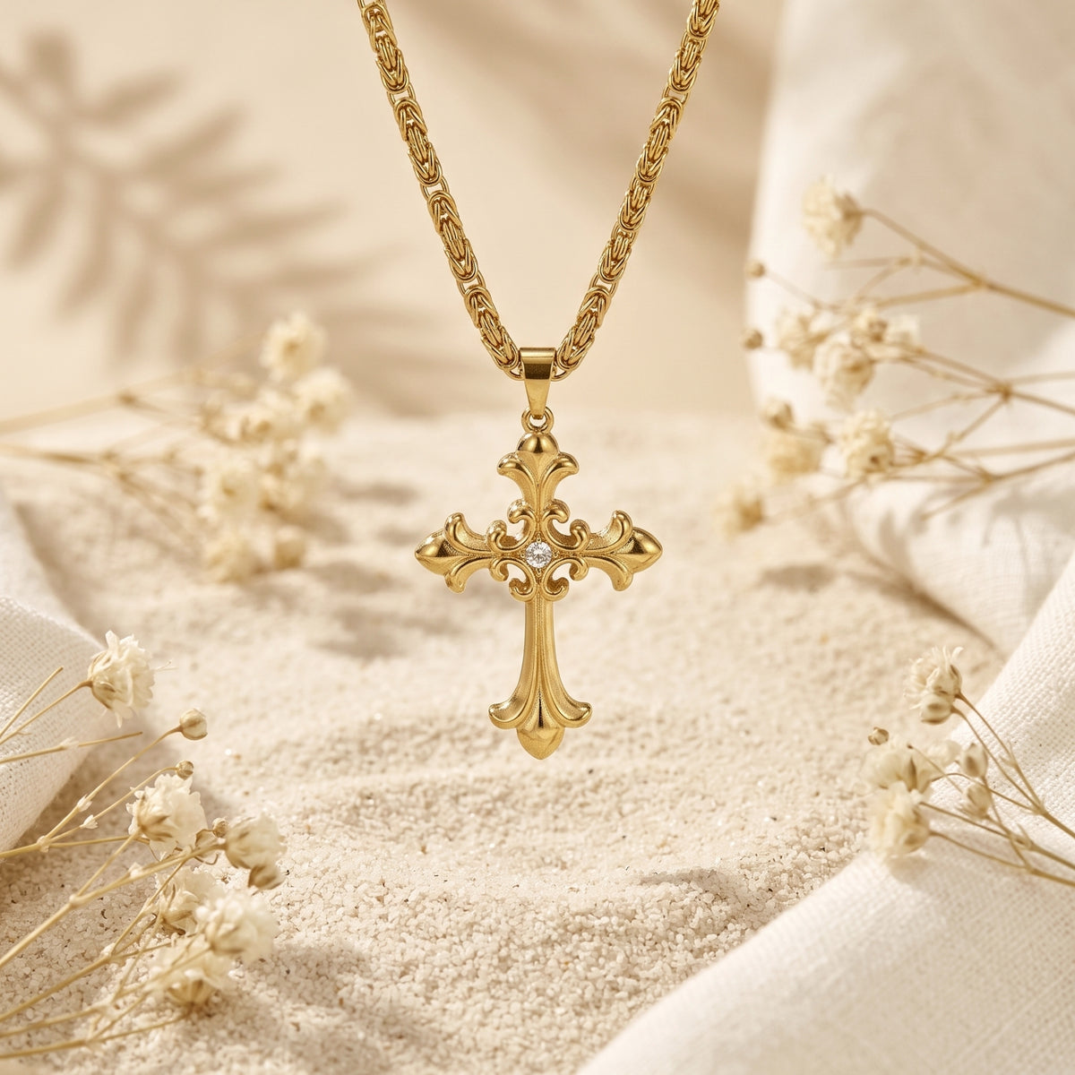 Front view of a gold-tone filigree cross pendant suspended upright over sand with dried white flowers.