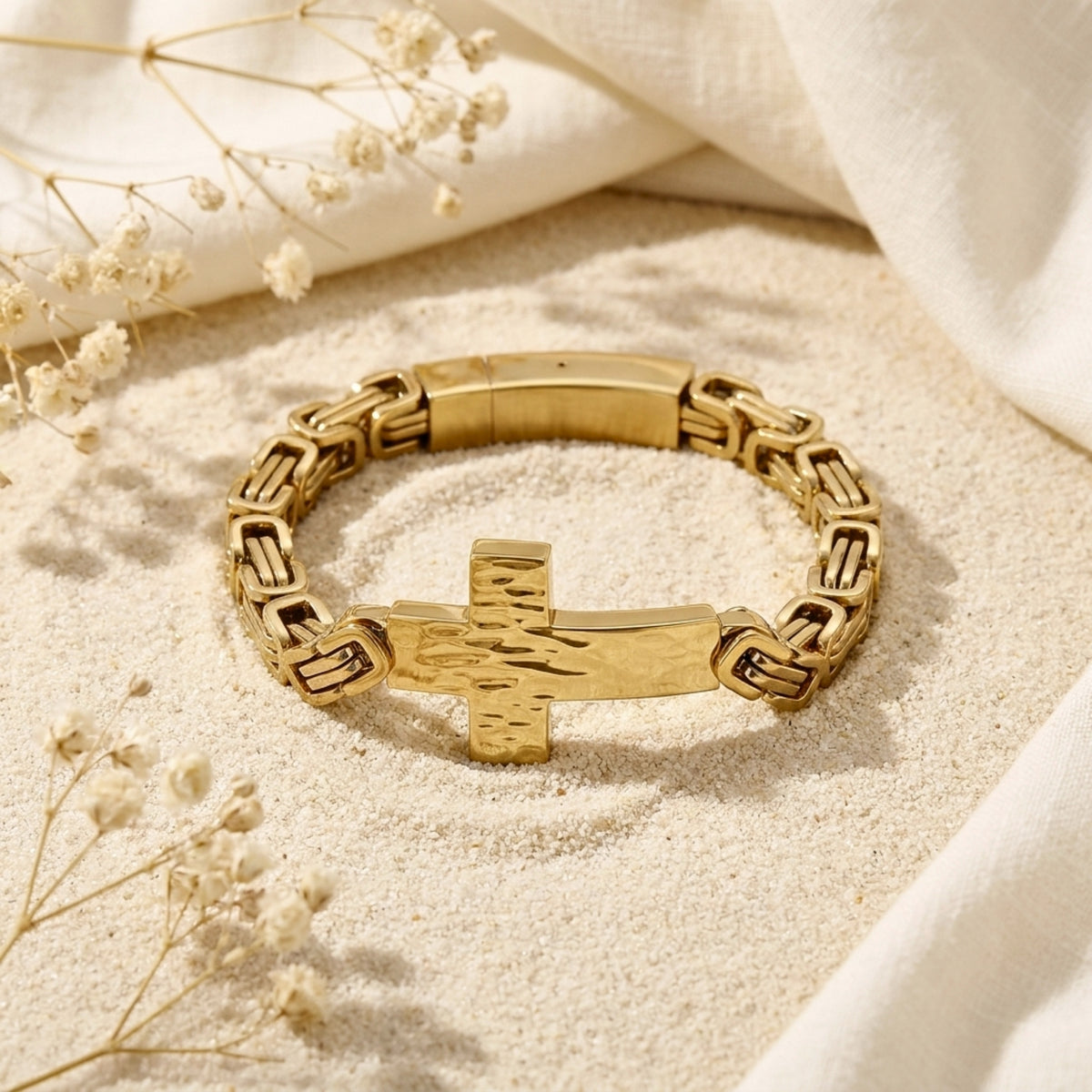 Gold-tone cross link bracelet displayed flat on sand, highlighting the hammered cross and foldover clasp.