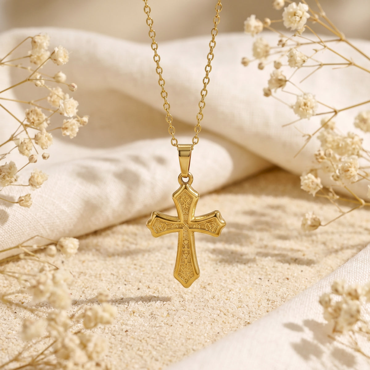 Gold-tone filigree cross pendant hanging vertically over sand and white fabric with dried flowers.
