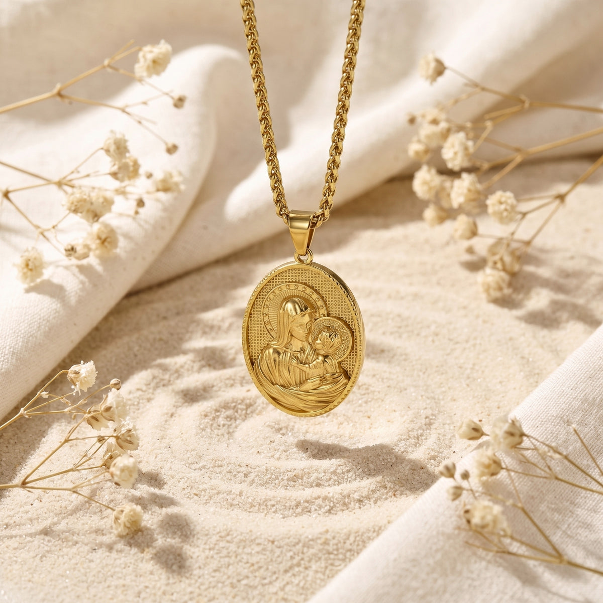 Gold-tone Madonna and Child medallion pendant suspended over sand among dried flowers and white fabric.