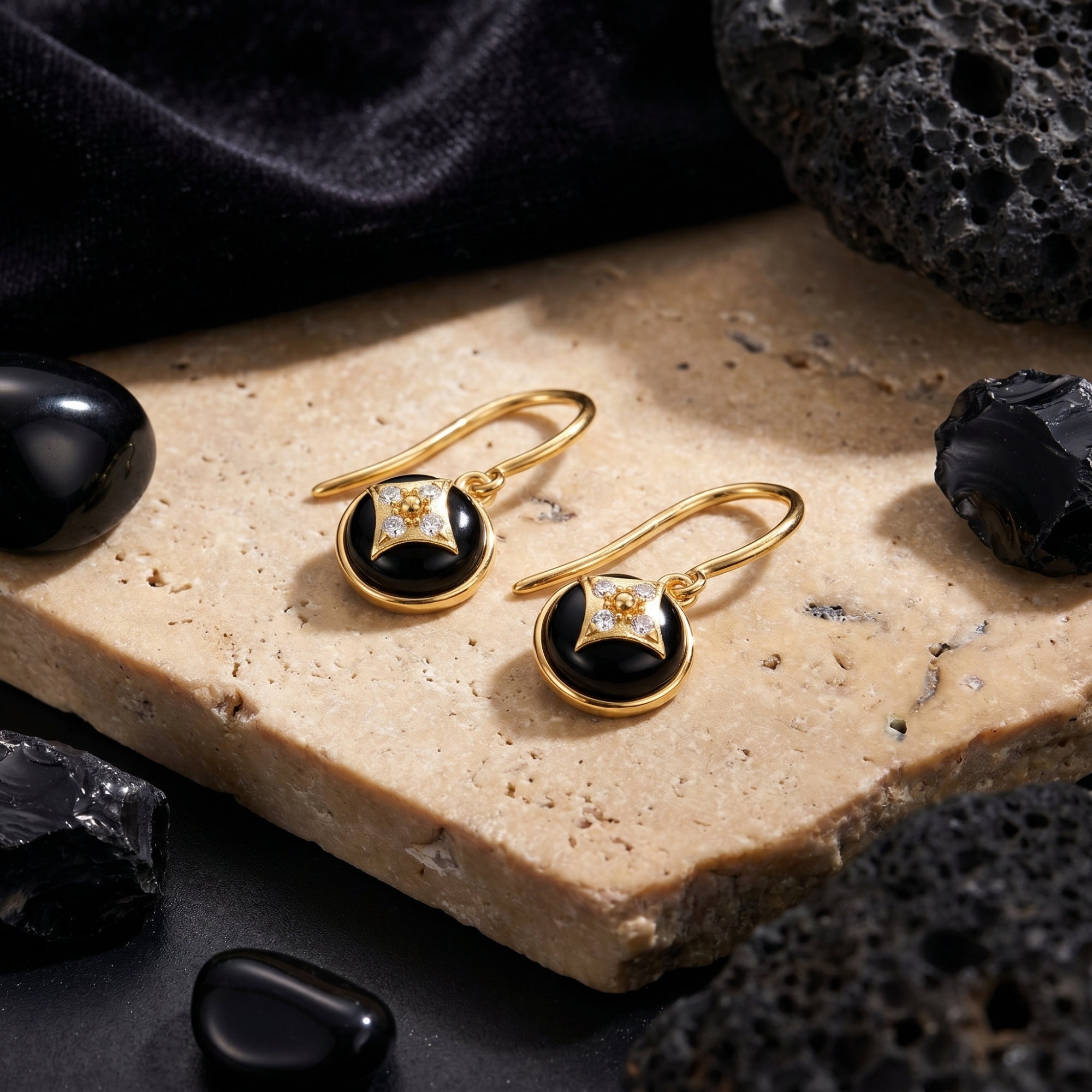 Pair of yellow gold drop earrings featuring round black onyx stones, styled on a stone slab with lava rocks.