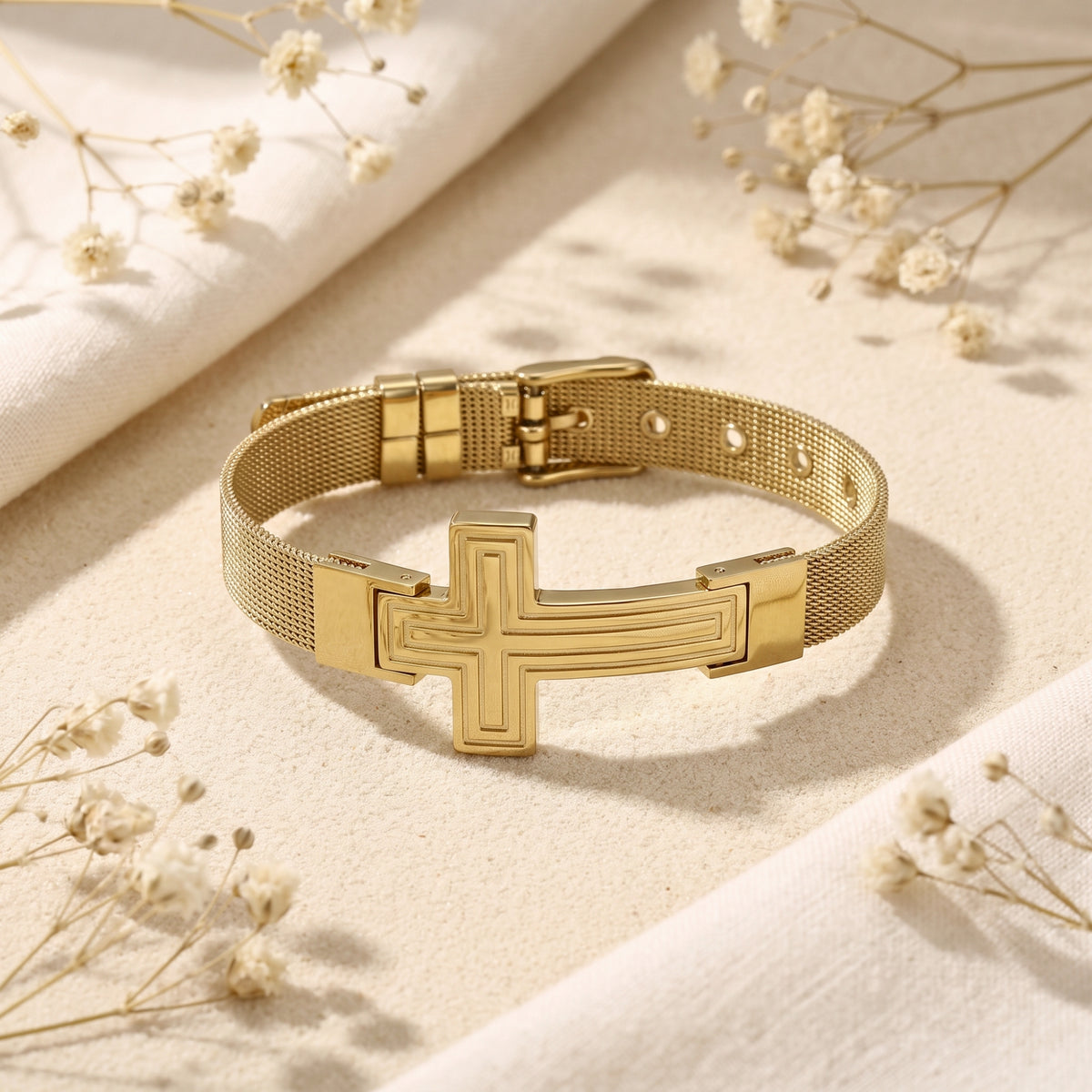 Gold-tone cross bracelet with an adjustable mesh strap and buckle closure, displayed flat on sand.
