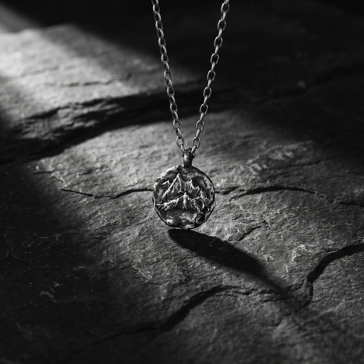 Silver mountain-relief medallion pendant suspended above dark slate, casting a shadow.