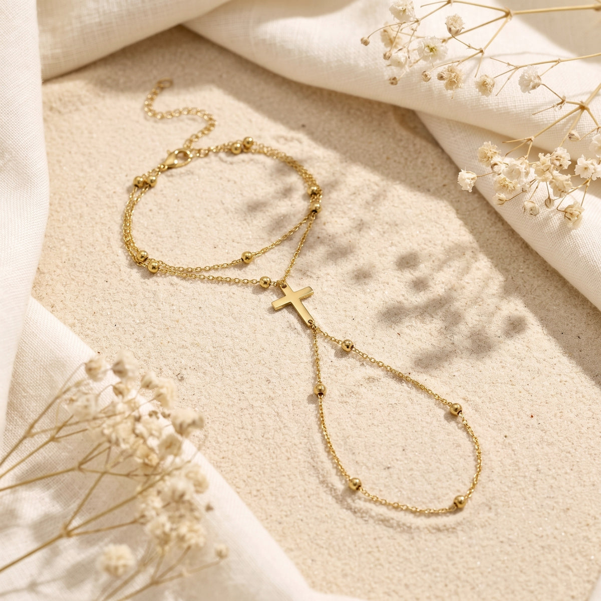 Flat lay view of the gold cross hand chain bracelet on textured sand framed by soft cream fabric.