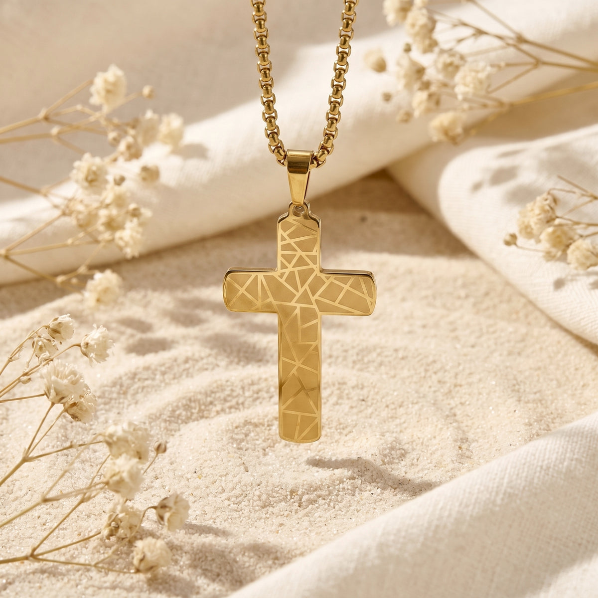 Gold geometric cross pendant suspended centrally over sand, surrounded by draped white fabric and dried flowers.