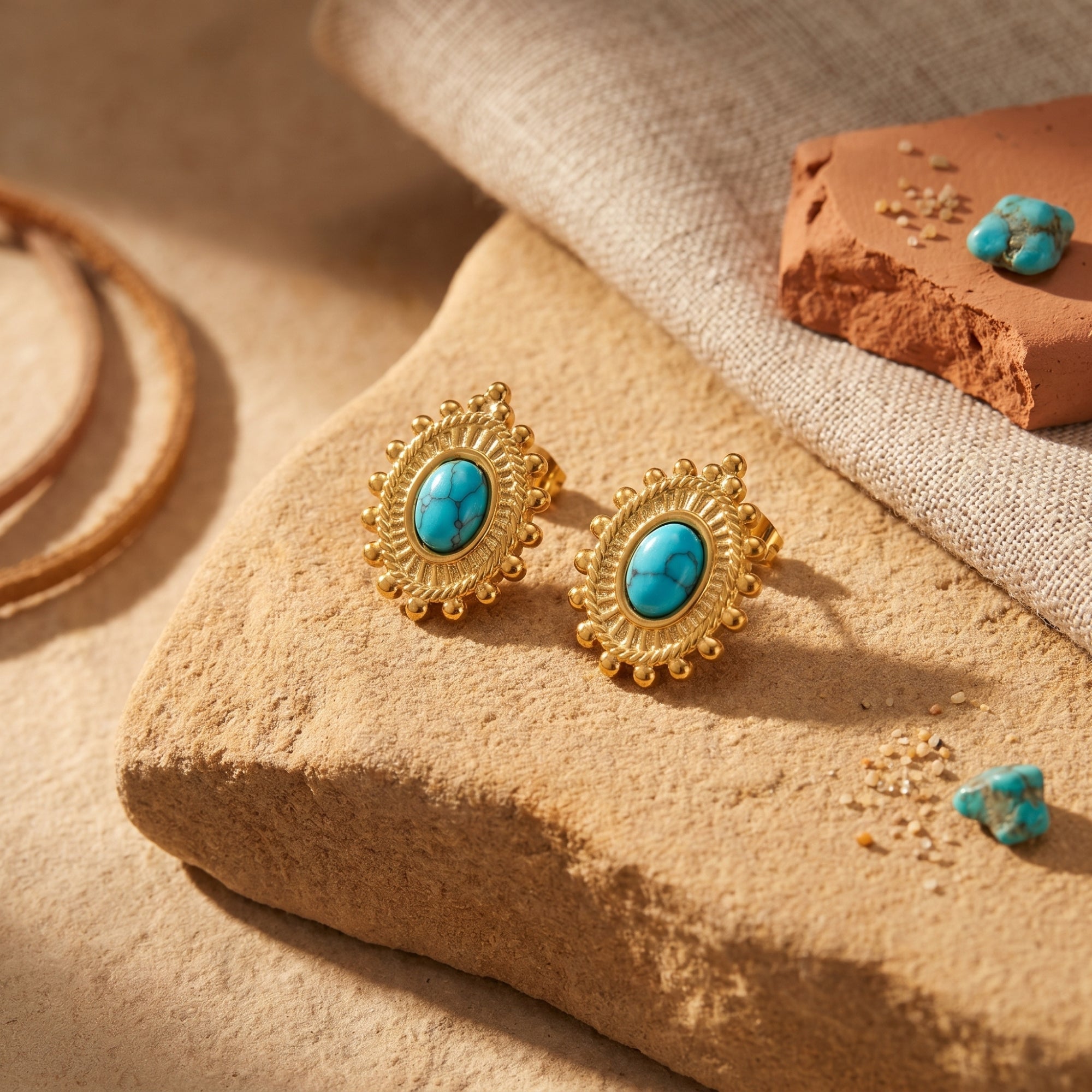 Pair of gold-tone turquoise-hued oval cabochon drop earrings displayed on a textured stone surface with fabric.