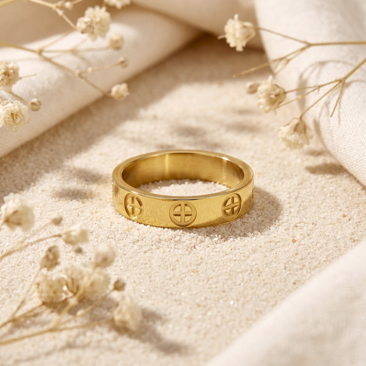 Front view of a gold-tone cross band ring lying on fine sand, with circular cross motifs and dried flowers around it.