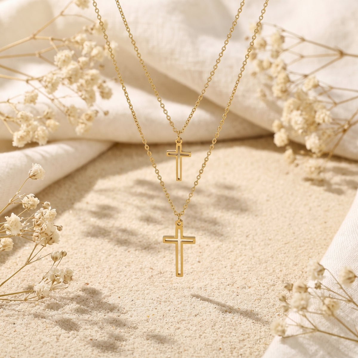 Gold-tone layered double cross necklace arranged on pale sand with dried flowers and white fabric in the background.