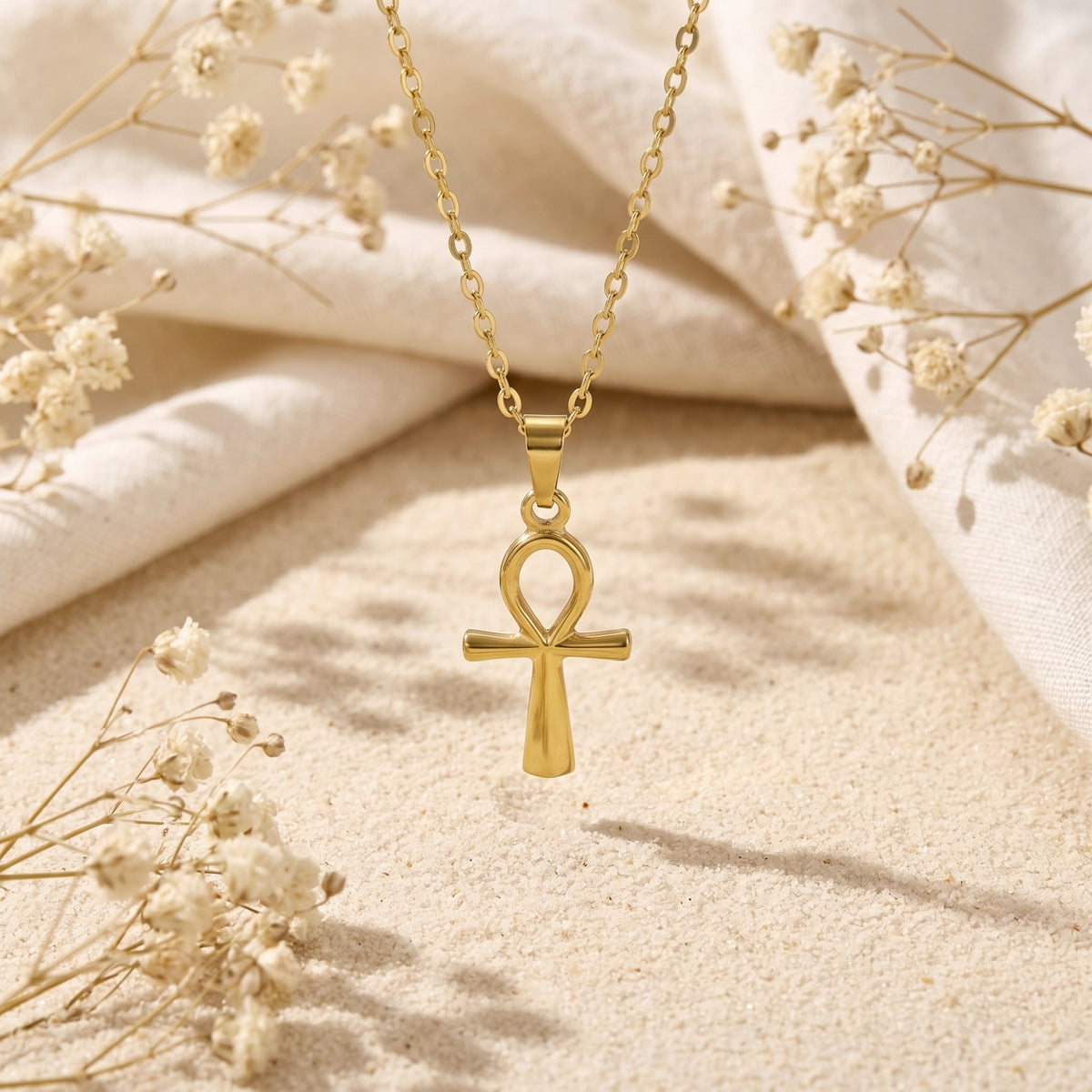 Gold-tone Ankh cross necklace displayed vertically over textured sand with fabric and dried flowers.