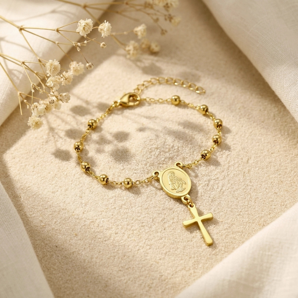 Angled view of a gold-tone rosary bracelet with round beads, Virgin Mary medal, and cross charm on beige fabric.