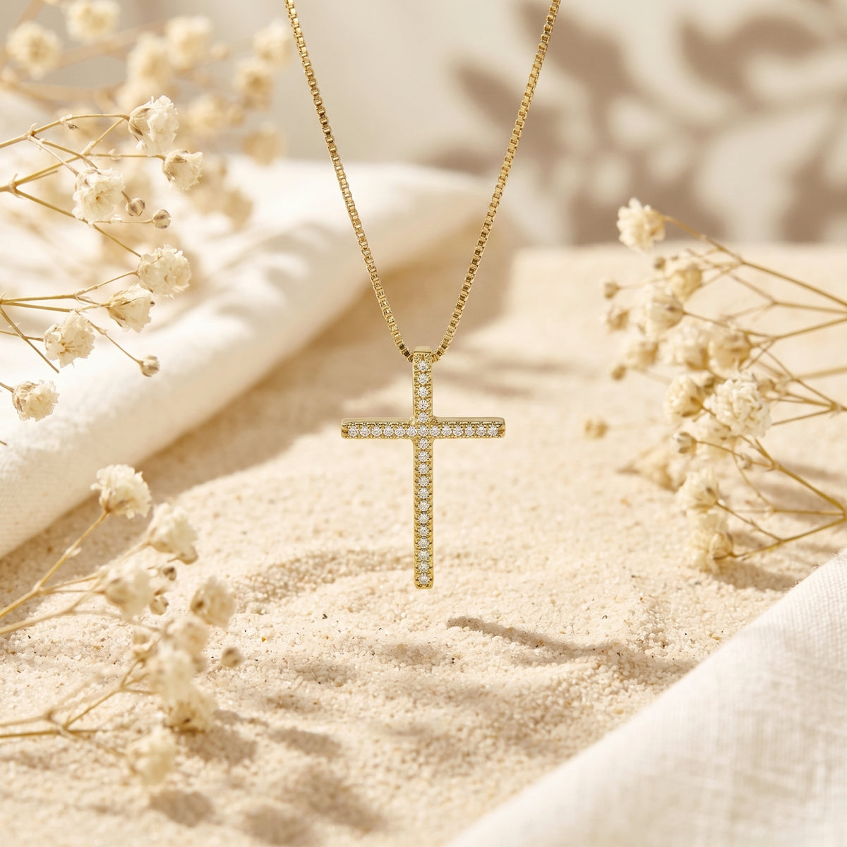 Gold-tone pavé cross necklace hanging above sand between white dried flowers.