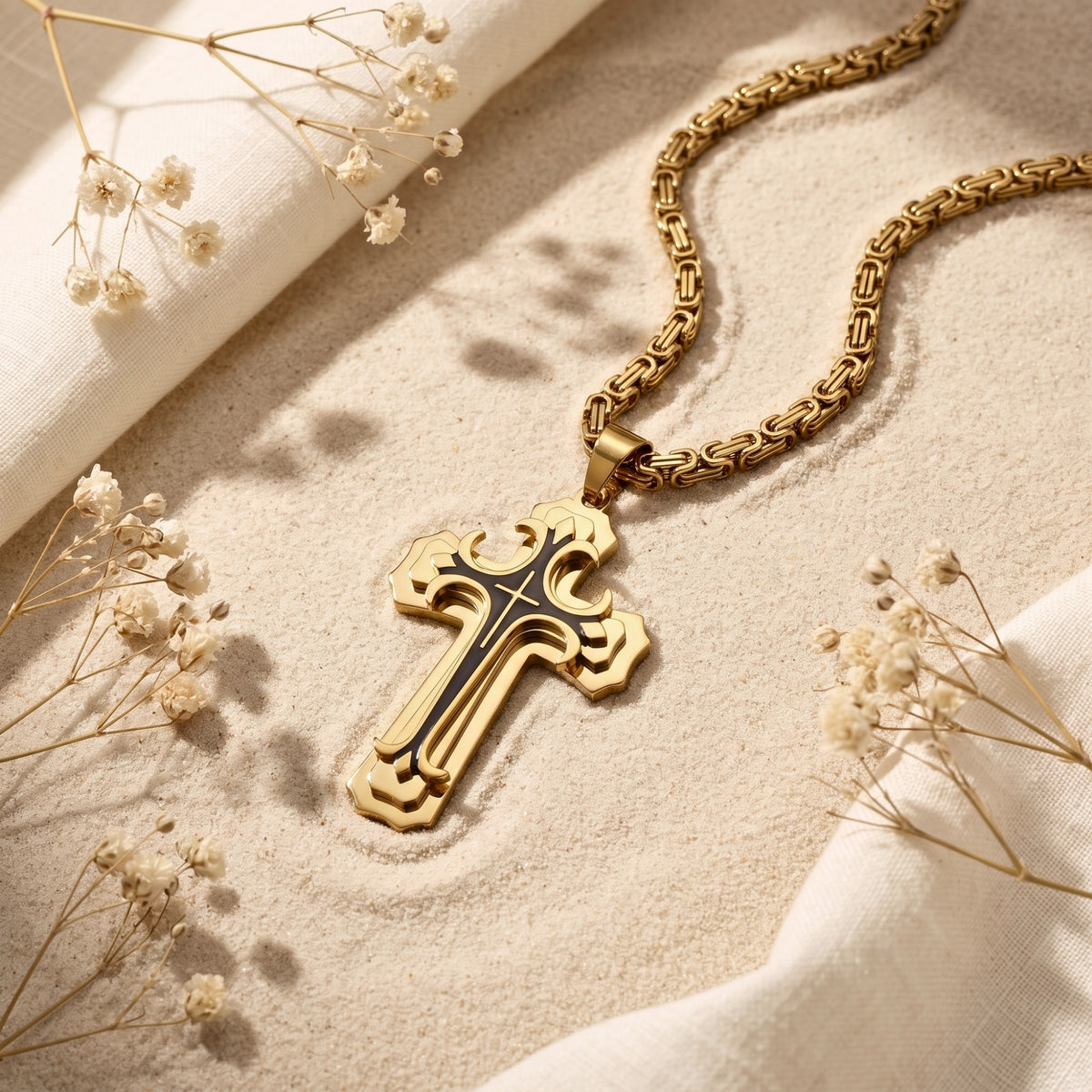 Gold-toned scroll cross pendant and chain lying diagonally on sand beside white fabric and dried flowers.