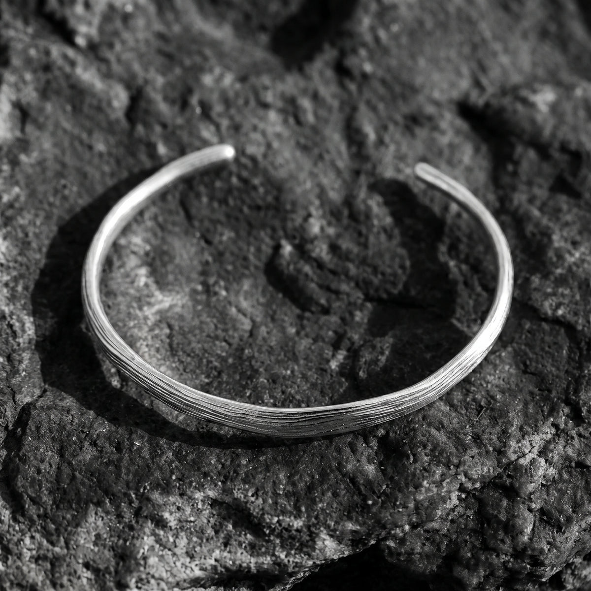 Top-down view of a textured silver open cuff bracelet resting on rough dark rock.