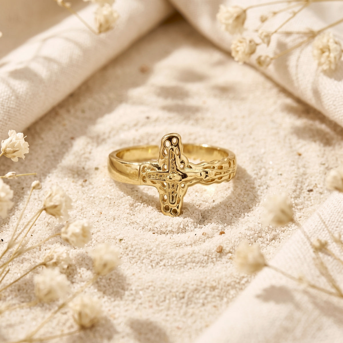 Front view of a gold-tone sculpted cross ring standing on sand between draped fabric.