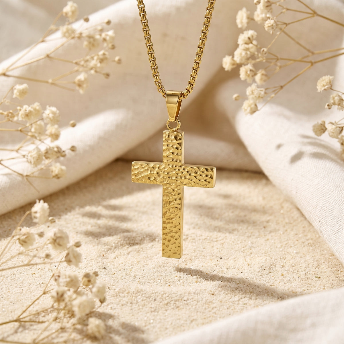 Gold-tone hammered cross pendant hanging upright over sand, with cream fabric and dried flowers in the background.