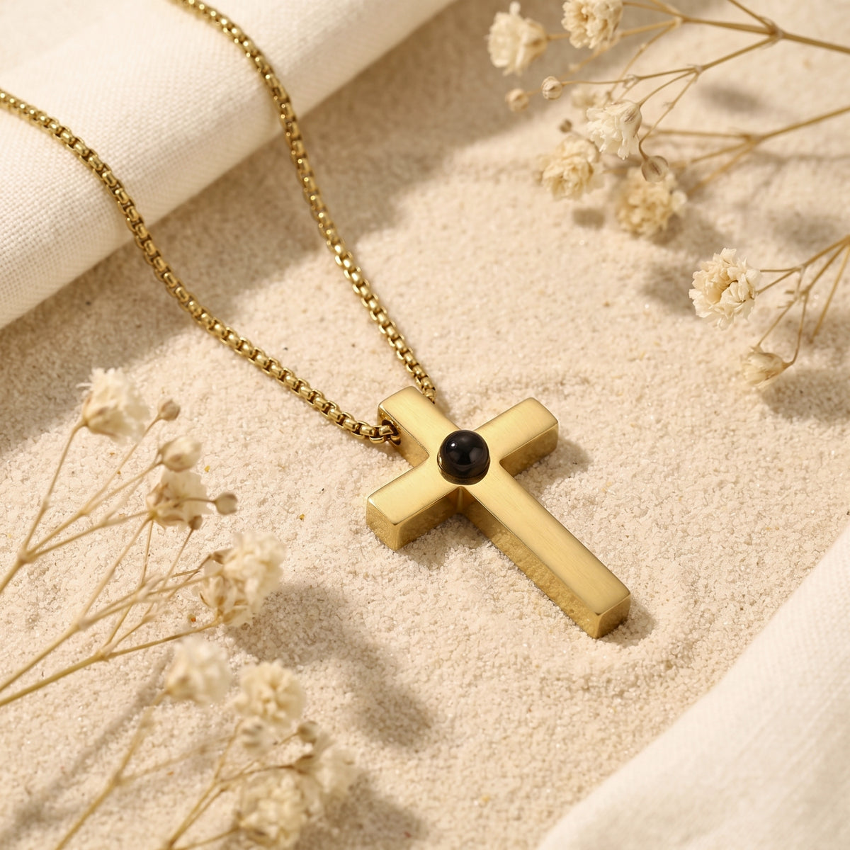 Gold-tone cross pendant with a central dark lens lying on sand among dried flowers.